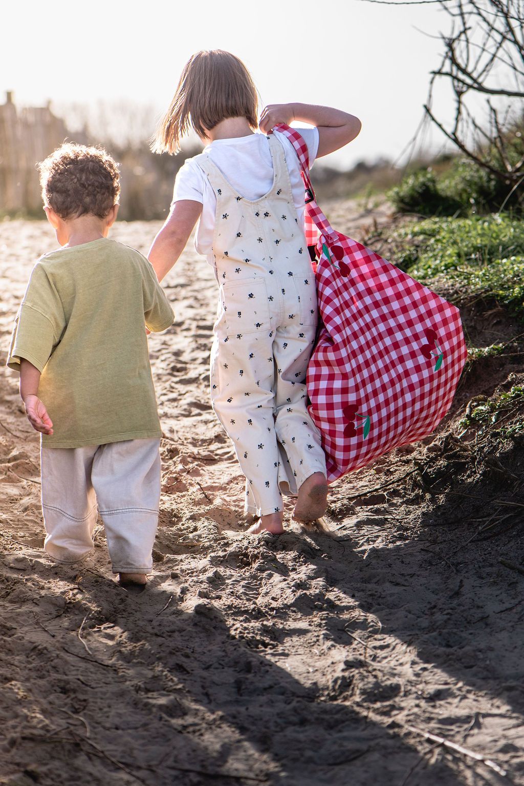 pic nic outdoor playmat, child is holding the closed bag on its shoulder while giving another child a hand, they walk in the sand